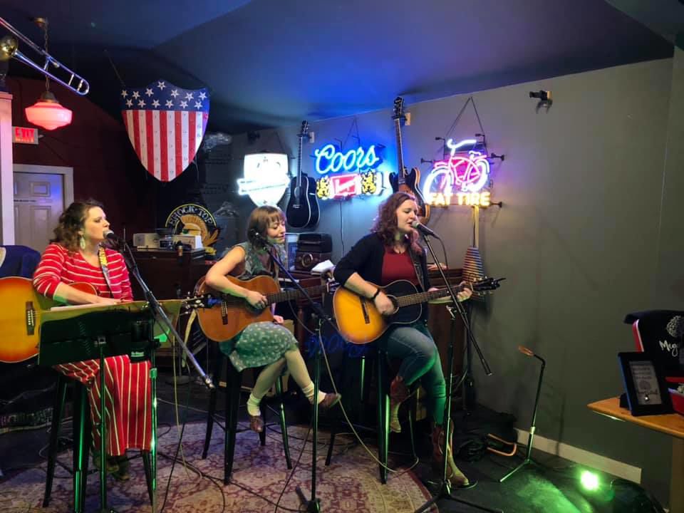 Three women sit while holding guitars as they sing into microphones